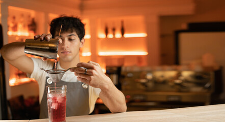 man bartender carefully filters bright alcoholic cocktail from steel shaker cup into glass through sieve. Close-up view.