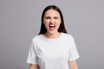 Screaming young beautiful adorable woman fed up with something looking at camera standing on grey background in studio isolated wearing casual white t-shirt angry sad desperate expressions.