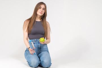 A young brown-haired girl holds a bottle of water and a yellow apple in her hands. Model wearing a gray top and blue jeans on a white background