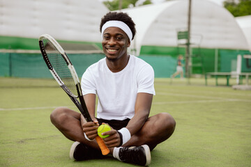 Man of African appearance is sitting on grass of tennis court, looking at the camera and smiling....