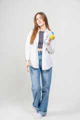 Beautiful young brown-haired girl in a white shirt and blue jeans holds a bottle of water and a yellow pear on a white background