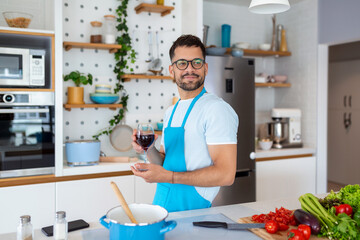 Home cook. Single man in apron looking at the window, drinking wine from a glass while cooking in the kitchen, cooking at home