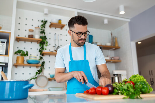 Happy young man chopping fresh vegetables or salad leaves, preparing vegetarian salad, enjoying cooking alone in modern kitchen standing at wooden countertop, hobby activity concept.