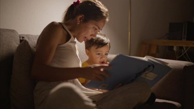 Cute latin 2 year toddler having a snack while his mother is reading a book at night while sitting on a couch