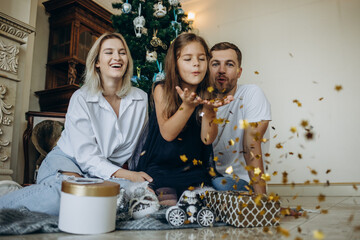 family of four parents pregnant mom and dad and two siblings little boy and girl in stylish clothes in chalet are ready to welcome Christmas and New Year near Christmas tree with gift box presents