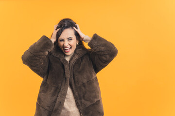 Young adult latin american woman wearing fur coat messing with her hair looking at camera with crazy laughing expression. Yellow background horizontal studio shot. High quality photo