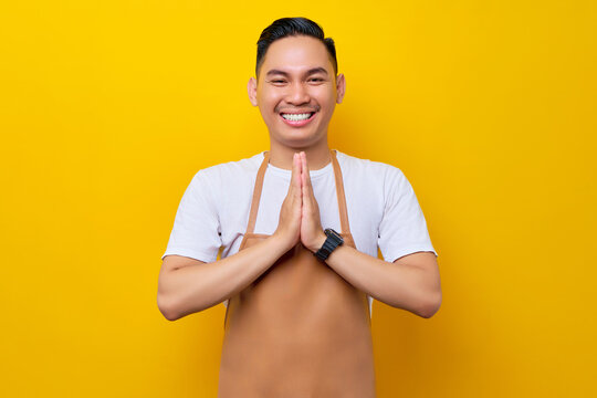 young Asian man 20s barista employee wearing brown apron working in coffee shop, holding hands together, greeting clients with smiling friendly on yellow background. Small business startup concept