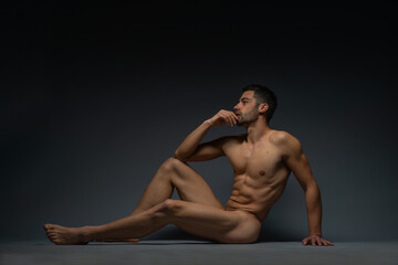 Fashion nude photo of a male model with seductive figure sitting isolated on the floor in a studio