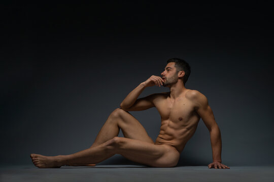 Fashion nude photo of a male model with seductive figure sitting isolated on the floor in a studio