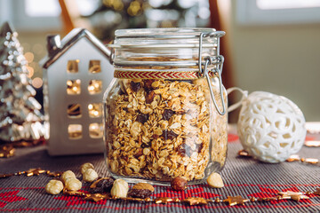 Concept of homemade Christmas present, handmade roasted granola with various nuts and raisins in beautiful glass jar. Blur Christmas tree and lights on background. - obrazy, fototapety, plakaty