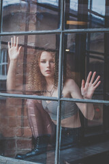 Beautiful attractive woman with curly hait and grey top touching window glass