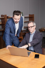 Mature businessman in suit is showing something on the laptop to pensive co-worker in the office