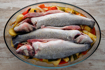 
Photo of three raw sea bass in a dish ready to bake with spices and vegetables on a wooden table. top view