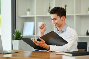 Fototapeta premium Happy asian male office worker checking financial reports and using laptop at his working desk