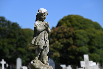 Old, worn, and weathered stone sculpture of a robed child looking down mournfully, hands clasped together in prayer, in a cemetery