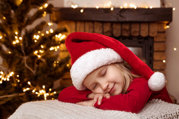 Fototapeta na wymiar Christmas Child Little Girl in Red Santa Hat is sleeping Waiting for Santa in Christmas Home Atmosphere by Fireplace and Tree. Christmad Holiday