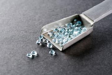 A handful of natural sky blue round topazes in a metal scoop of tweezers. Dark gray paper background. Natural topaz small round faceted gem stone settings for making jewelry from gold or silver. 