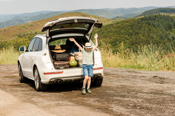 Child stands next to the full trunk of an SUV. Child comes with us on vacation and it is driven by...