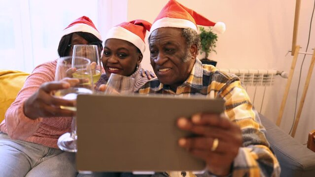 Happy African American family of three making a video call with a tablet, white wine and Santa hats celebrating Christmas. Focus on daughter