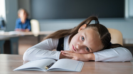 Little caucasian girl is bored at the lesson at school. The schoolgirl folded her head on the desk and the teacher sits in the background. 