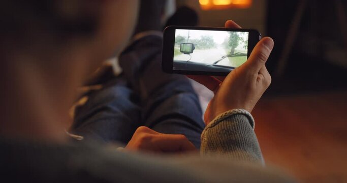 Hands, phone and man with dash cam video, screen and display relax on a sofa, streaming travel vlog in his home. Behind, smartphone and ui with guy chilling in a living room, watch show in winter