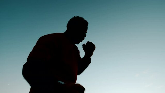 Bottom view of a silhouette black man in sportswear and bondage practicing boxing by conducting shadow fight against background sky outdoors sunset