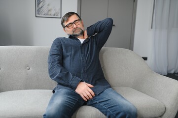 Fototapeta premium Neck Pain. Portrait Of Exhausted Mature Man Massaging Aching Neck Suffering From Ache Sitting On Couch At Home In Living Room. Osteoarthritis, Older People Health Problem, Sedentary Lifestyle Concept
