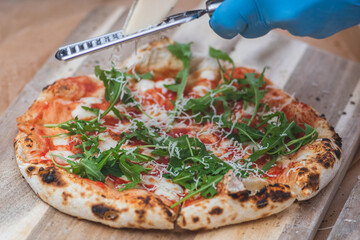Hand with plastic glove grating with a grater parmesan cheese on a pizza with tomato sauce,...