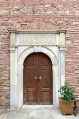 Fototapeta na wymiar White marble decorated entrance of Arap Mosque, with wooden arched door, in red stone bricks wall, Karakoy, Beyoglu district, Istanbul, Turkey. Text above door says: Enter here in peace, secure