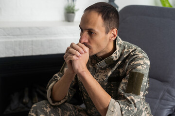 Thoughtful military man staring aside, holding palms by mouth, sitting on couch at home. Young soldier visiting psychologist, suffering from posttraumatic stress, closeup photo, copy space.