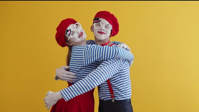 A male mime and a female mime in striped and bright suits smile and hug tightly. Pantomimes act out a performance on a yellow background