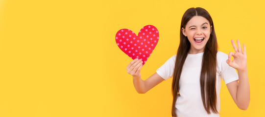 winking kid hold love heart show ok gesture on yellow background, february 14. Kid girl portrait with heart love symbol, horizontal poster. Banner header with copy space.