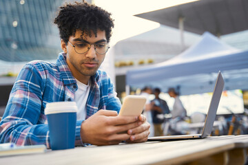 Curly haired middle eastern man holding mobile phone, reading text message in cafe. Pensive successful freelancer using smartphone, laptop working online sitting at workplace. Technology concept 