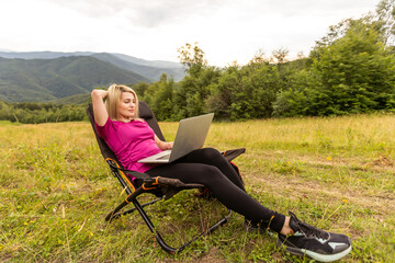 A woman in a sports warm suit works on a laptop outdoors in a mountainous area.
