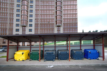 Fototapeta premium New colored plastic garbage bin for sorting various household waste. Colorful plastic bins for different waste under conopy next to new residential building. Place for dumpsters to collect garbage