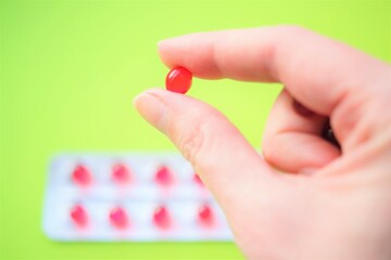 Round red vitamin in human fingers. Blurred new blister with capsules on green table