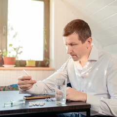 A sick person feels the symptoms of a viral disease, holds a thermometer in his hand. Tablets and water on the table.