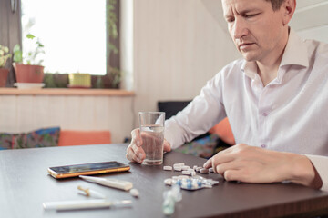 A sick person feels the symptoms of a viral disease, holds a thermometer in his hand. Tablets and water on the table.