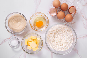 Basic baking ingredients in clear glass bowls to include eggs, flour, sugar, butter, salt with copy space
