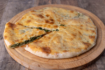 Pie with spinach, herbs and suluguni cheese on a wooden board. Whole pie with cut off piece. Traditional round flat pie with filling. Homemade of Caucasian and Balkarian cuisine
