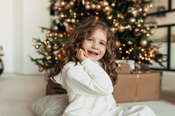Lovely cute adorable little girl with curls wearing white dress sitting in front of Christmas tree with happy smile 
