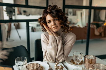 Charming pretty lady with curly hair wearing sweater posing at camera while sitting on holiday dinner table. 