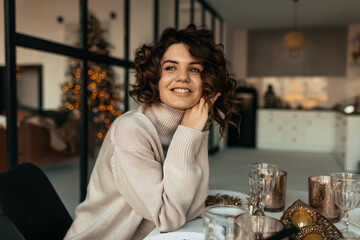 Charming pretty lady with curly hair wearing sweater sitting on dinner holiday table and looking at window with smile