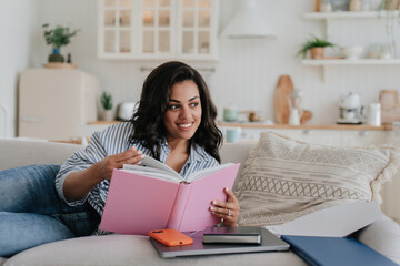 Cheerful African American young woman in striped shirt and blue jeans laying on sofa with opened...