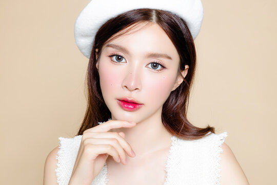 Cute Asian woman with perfect clear fresh skin. Pretty girl model wearing white beret and natural makeup on beige background. Cosmetology, beauty and spa, wellness, Plastic surgery.