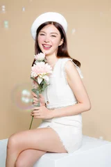 Fotobehang Cute Asian woman with perfect skin and holding pastel flower. Pretty girl model wearing white beret and natural makeup on beige background. Cosmetology, beauty and spa, wellness, Plastic surgery. © Auttapol