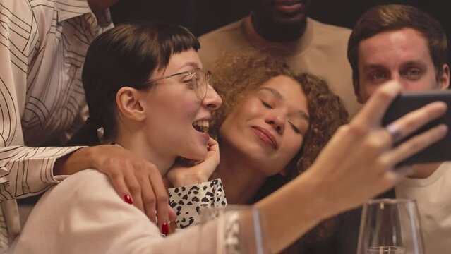 Group of joyful young diverse friends in fancy outfits recording funny videos and taking selfies on smartphone camera while enjoying time together at dinner party