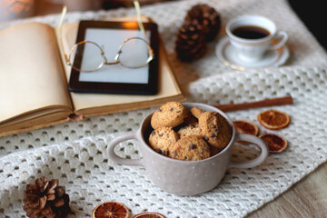 Cup of tea, books, tablet, bowl of cookies, various spices, pine cones and lit candles. Hygge at home. Selective focus.