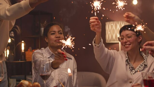 Group of young happy diverse friends burning sparklers during Christmas dinner party in cozy domestic atmosphere