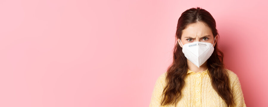 Preventive Measures, Health Care Concept. Close Up Of Angry Woman In Respirator Frowning, Looking With Judgemental Face At Person Without Mask, Standing Against Pink Background